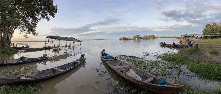 Fotografía panorámica del río Loretayacu bajo un cielo nublado al amanecer. En primer plano, varias canoas de madera largas y estrechas (peque-peques) están atracadas en la orilla lodosa; algunas tienen motores fuera de borda y otras contienen redes o suministros cubiertos con lonas azules. A la izquierda, una estructura techada sencilla se levanta sobre el agua. A la derecha, un grupo de personas se congrega cerca de las embarcaciones en una zona de pasto verde. Al fondo, se observan casas flotantes de colores y una densa línea de vegetación selvática en el horizonte.
