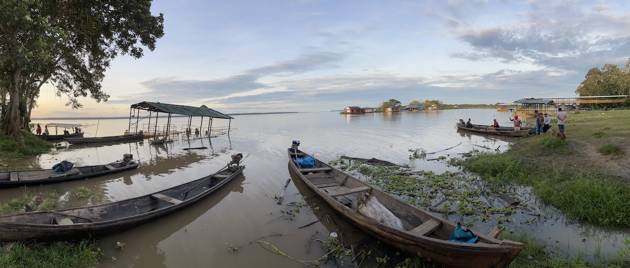 Fotografía panorámica del río Loretayacu bajo un cielo nublado al amanecer. En primer plano, varias canoas de madera largas y estrechas (peque-peques) están atracadas en la orilla lodosa; algunas tienen motores fuera de borda y otras contienen redes o suministros cubiertos con lonas azules. A la izquierda, una estructura techada sencilla se levanta sobre el agua. A la derecha, un grupo de personas se congrega cerca de las embarcaciones en una zona de pasto verde. Al fondo, se observan casas flotantes de colores y una densa línea de vegetación selvática en el horizonte.