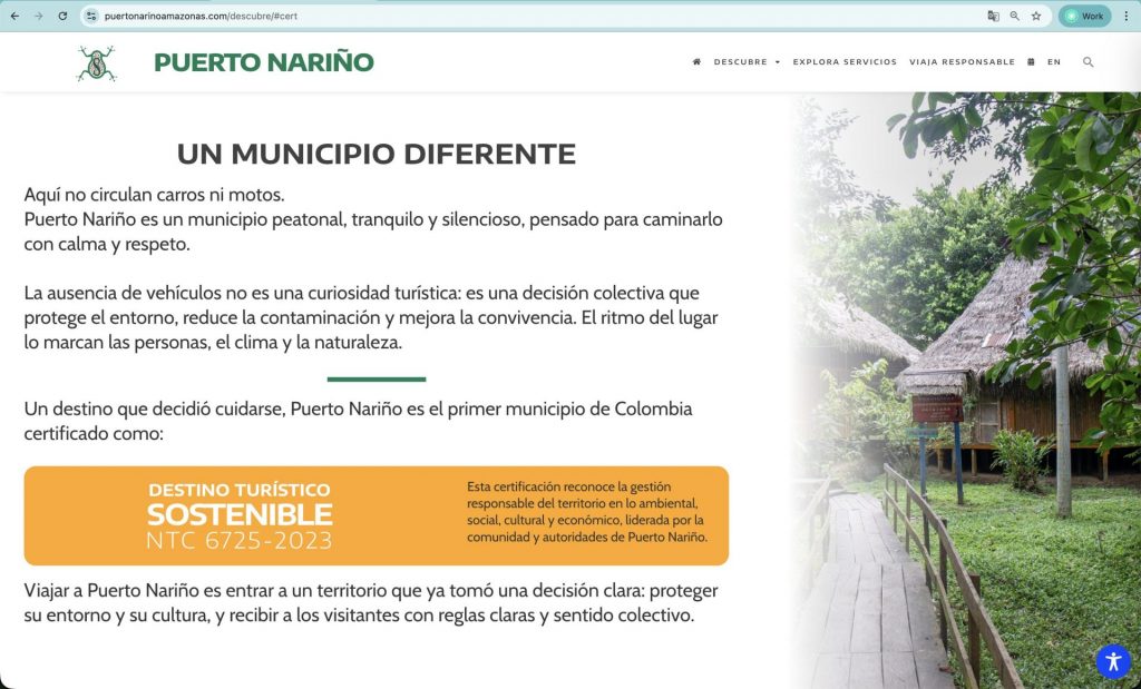 Captura de pantalla de la sección "UN MUNICIPIO DIFERENTE" del sitio web de Puerto Nariño, Amazonas. El texto describe al municipio como un lugar peatonal donde no circulan carros ni motos por decisión colectiva para proteger el entorno. En la parte central destaca un recuadro naranja que indica que es el primer municipio de Colombia certificado como "DESTINO TURÍSTICO SOSTENIBLE NTC 6725-2023". A la derecha, se muestra una fotografía de un sendero peatonal de madera que conduce hacia malocas tradicionales rodeadas de abundante vegetación selvática. La interfaz mantiene el logo de la rana verde y el menú de navegación superior.