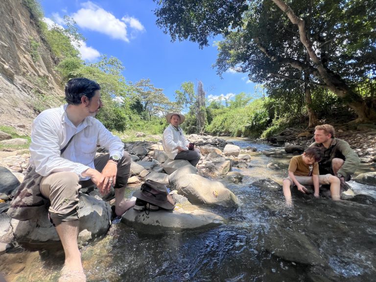 Un grupo de cuatro personas, tres adultos y un niño, descansa junto a un río poco profundo y pedregoso en un entorno natural y boscoso bajo un cielo azul con nubes dispersas. En primer plano a la izquierda, un hombre con camisa blanca y pantalones caqui está sentado descalzo sobre una roca grande, mirando hacia el centro. En el medio, otro hombre con sombrero de safari y camisa clara se sienta sobre las rocas del río. A la derecha, un hombre y un niño pequeño están sentados directamente en el agua fresca del río, jugando con las piedras. El paisaje está rodeado de vegetación densa, árboles altos y una ladera empinada a la izquierda.